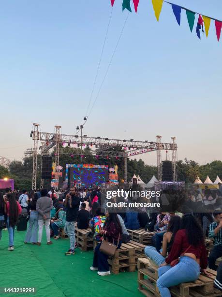 image of people milling around horn ok please food festival exhibition grounds with fake grass, metal construction frame pergola covered with multi-coloured streamers, large screen performance stage, focus on foreground - friends tv show stock pictures, royalty-free photos & images