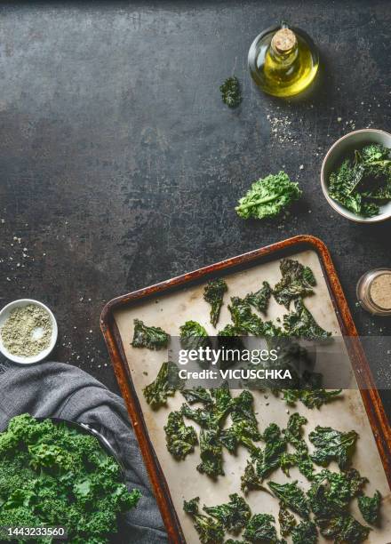 baked kale chips on baking sheet on dark kitchen table with ingredients: olive oil and herbs salt . - kale chips fotografías e imágenes de stock