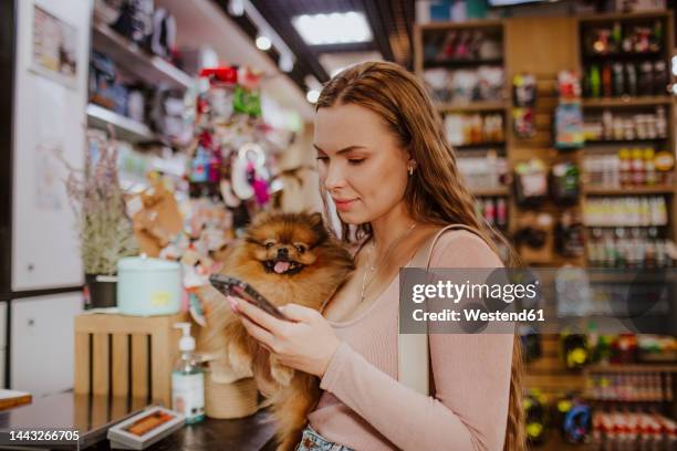 woman carrying dog using smart phone in pet shop - pet shop stock pictures, royalty-free photos & images