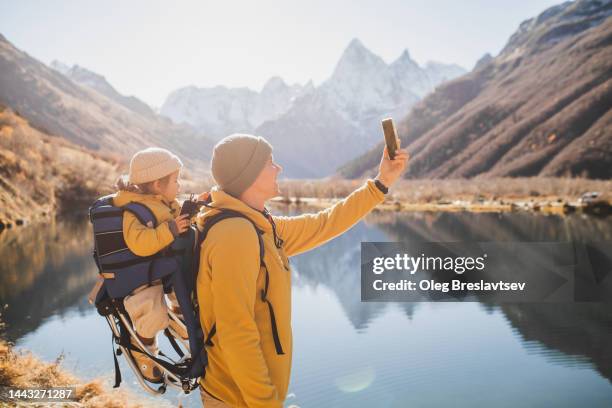 father with child in baby carrier backpack hiking in mountains and photographing with phone - vida sencilla fotografías e imágenes de stock