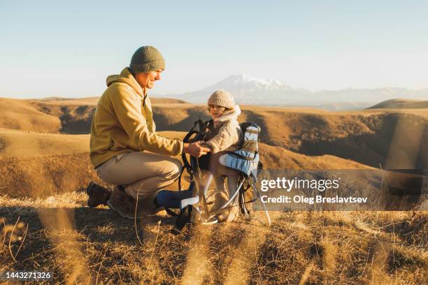 father seating little daughter in baby carrier backpack. meadow with mountain view at sunset - montagnes du caucase photos et images de collection