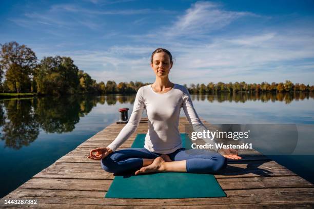 woman with eyes closed practicing meditation in front of lake - schneidersitz stock-fotos und bilder