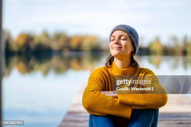 smiling woman with arms crossed and eyes closed sitting on jetty - mindfulness stock pictures, royalty-free photos & images