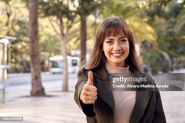 front view of a young caucasian woman outdoors in the city smiling and making thumbs up gesture with one finger of hand while looking at camera. - woman thumb stock pictures, royalty-free photos & images