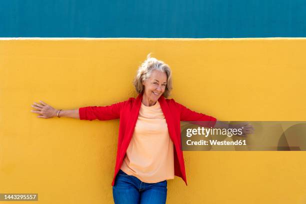 smiling woman with arms outstretched leaning on yellow wall - chaqueta roja fotografías e imágenes de stock