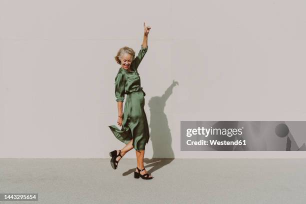 happy woman dancing with hand raised on footpath - older women stockfoto's en -beelden