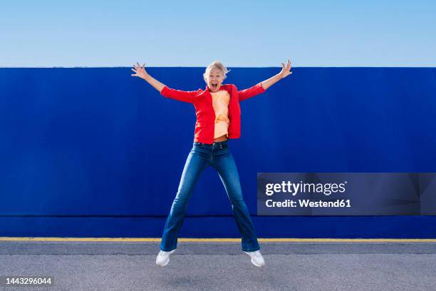 cheerful mature woman with arms outstretched jumping on footpath - frau arme hoch ganzkörper stock-fotos und bilder