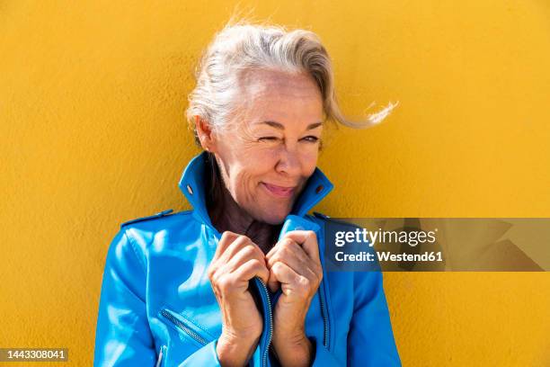 happy mature woman winking in front of yellow wall - winking stockfoto's en -beelden