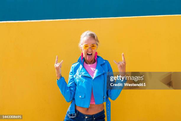mature woman sticking out tongue in front of colored wall - grey hair cool woman stock-fotos und bilder
