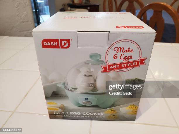 Eye-level shot of a 'DASH Rapid Egg Cooker' box with an image of whole eggs in a cooker, resting on a white tile countertop with wooden chairs in the...