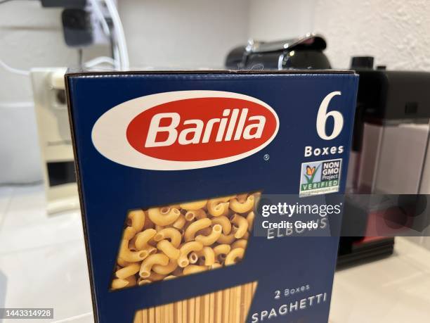 Eye-level shot of a bulk-buy 'Barilla' variety box, containing elbows, spaghetti, and penne resting on a white tile countertop with kitchen gadgets...