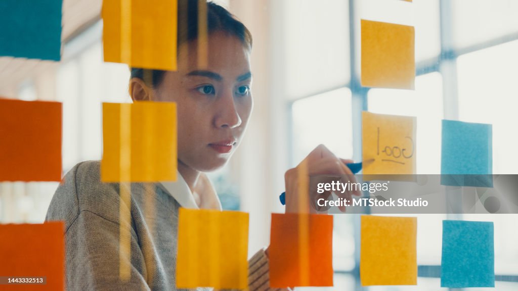 Asian girl focus on transparent board writing roadmap planning on sticky note work outside office in co-working room.