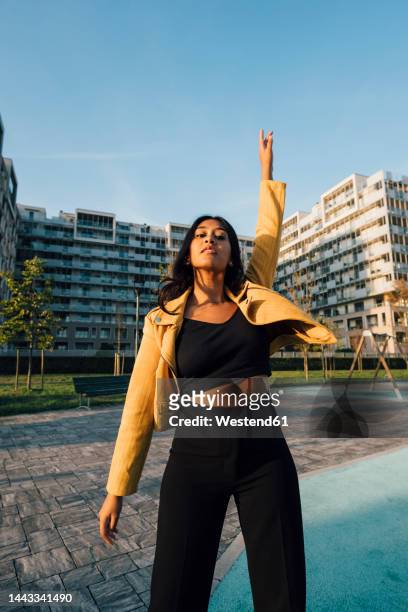young woman with arm raised standing in front of buildings at sunset - mains en l'air photos et images de collection
