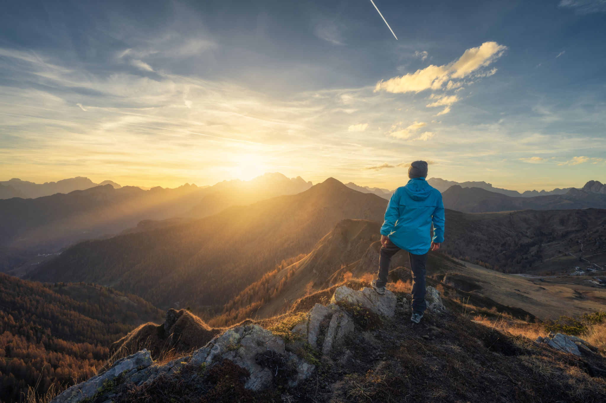 Man on stone on the hill and beautiful mountains in haze at colorful sunset in autumn. Dolomites, Italy. Sporty guy, mountain ridges in fog, orange grass and trees, blue sky with sun in fall. Hiking Man on stone on the hill and beautiful mountains in haze at colorful sunset in autumn. Dolomites, Italy. Sporty guy, mountain ridges in fog, orange grass and trees, blue sky with sun in fall. Hiking