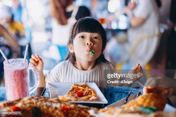 portrait of a playful little asian girl eating a piece of basil leaf and making funny face while having pizza margherita in an outdoor restaurant. having a fun time dining out with her family. family concept. family eating out lifestyle - restaurant kids stock-fotos und bilder