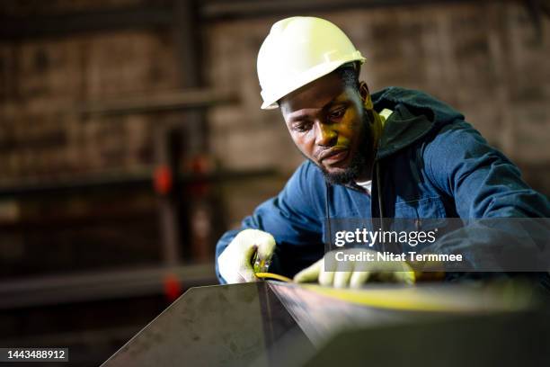 metalworking measuring tools for precision of a metal fabrication. a male african american industrial worker measuring the accuracy of a construction beam with a stainless measuring tape in a metal factory. - fabbro ferraio foto e immagini stock
