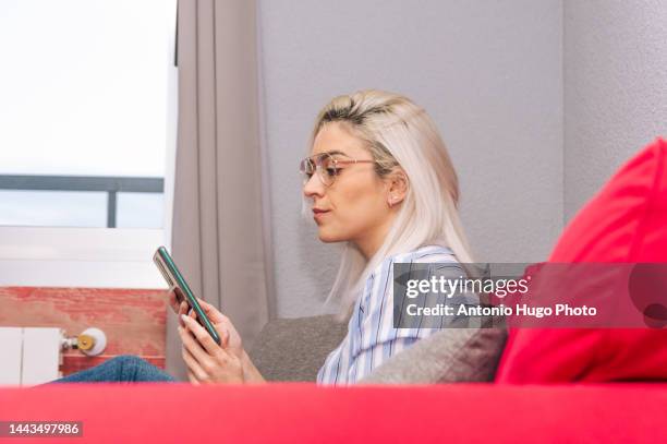 young blonde woman with platinum hair and eyeglasses dressed with a striped shirt using her mobile phone. sitting on the couch. relax time. - addiction mobile and laptop stockfoto's en -beelden