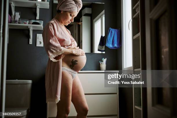 standing pregnant woman applying cream on her tummy - woman in shower tattoo stock pictures, royalty-free photos & images