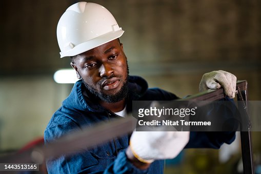 Welded Seam Inspection in Metal Structure Fabrication. Front view of a Male African American welder checking welded seam accuracy of a metal frame parts surface during welds in metal fabrication.