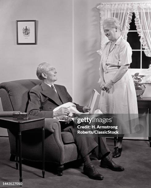 1940s 1950s Senior Man Sitting In Easy Chair Opening A Gift From His Wife Standing Before Him Smiling