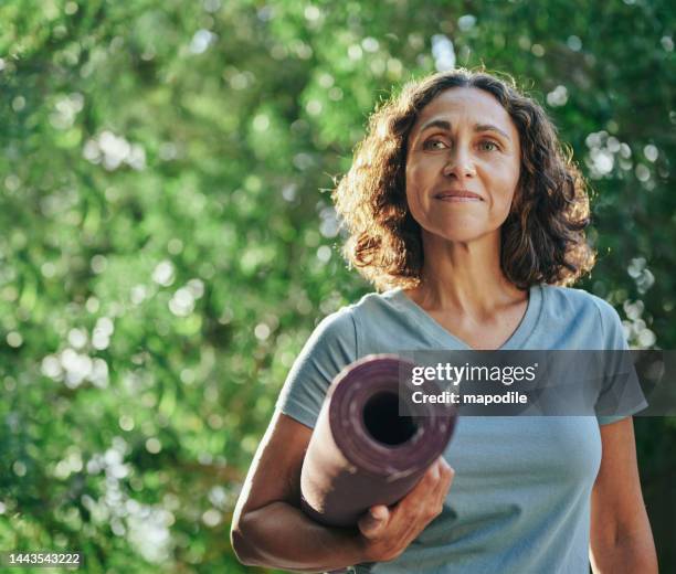 femme mûre souriante portant un tapis d’exercice debout dans un parc - tapis de sol photos et images de collection