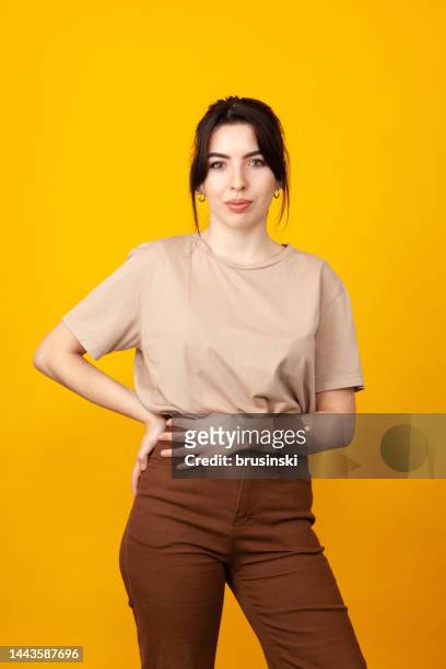 studio portrait of a cheerful 20 year old white woman with black hair in a beige t-shirt and brown pants against a yellow background - white t shirt studio imagens e fotografias de stock