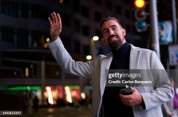 elegant caucasian guy at the city during the night time, raising an arm to ask for a taxi. businessman standing by the street hailing a cab. - uber taxi app in madrid stock pictures, royalty-free photos & images