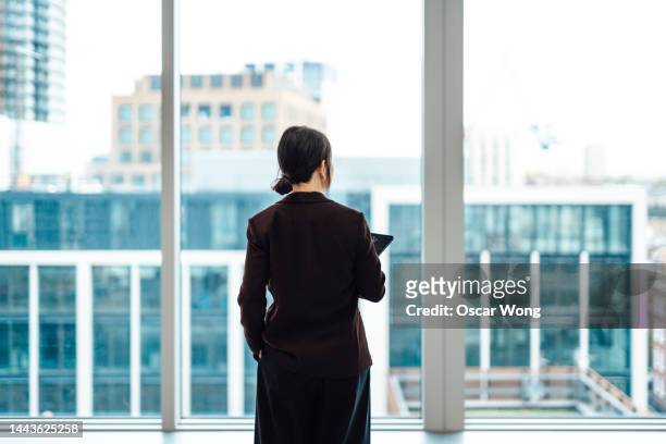 young asian businesswoman with digital tablet, standing against office window with city view - business woman rear view stock pictures, royalty-free photos & images