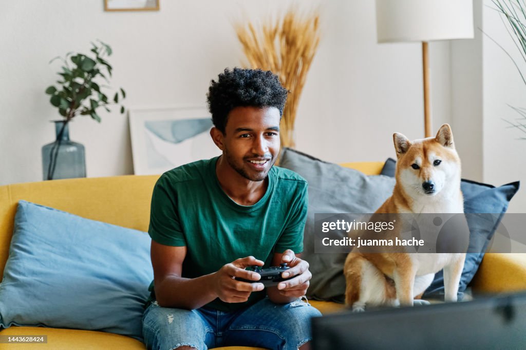 Man sitting on living room sofa at home playing video games accompanied by his dog sitting next to him.