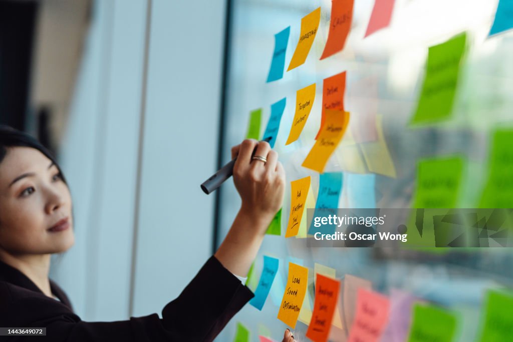Young Asian businesswoman brainstorming strategy and new ideas on glass wall with adhesive notes