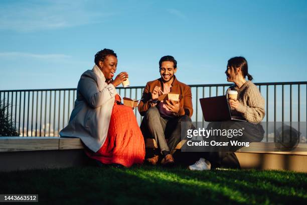 cheerful multi-ethnic business people sitting and having takeaway lunch at rooftop garden at top of office building during lunch break - smart casual lunch stock pictures, royalty-free photos & images