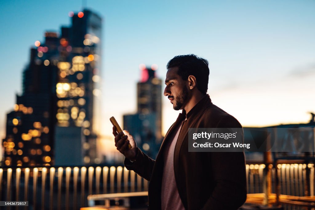 Businessman using mobile phone at rooftop in business building against illuminated financial buildings and London cityscape at night