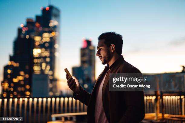 businessman using mobile phone at rooftop in business building against illuminated financial buildings and london cityscape at night - azioni e partecipazioni foto e immagini stock