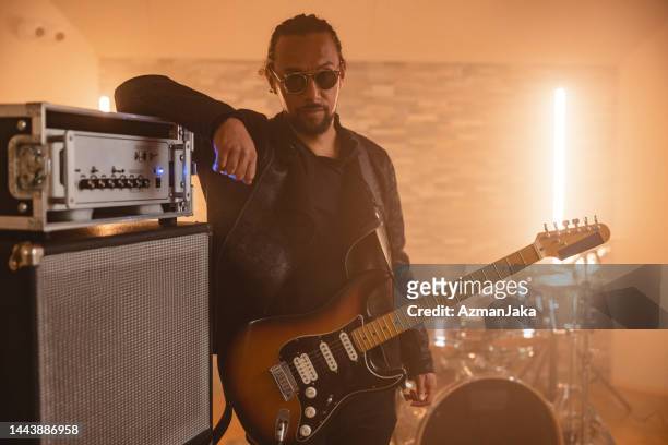 cocky black male musician with a guitar around his neck leaning against the music equipment in a small cosy home recording studio - amp stock pictures, royalty-free photos & images