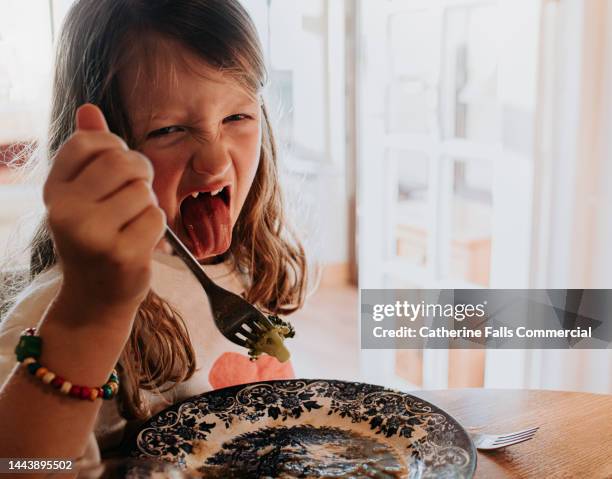 a little girl is disgusted by the broccoli on her fork. she sticks out her tongue to display her annoyance. - taste test stock pictures, royalty-free photos & images