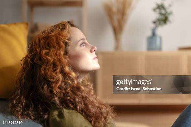 close up view of a young redhead woman sitting on the floor, leaning her back on sofa and looking into the distance with sad face. - young woman close at home stock pictures, royalty-free photos & images