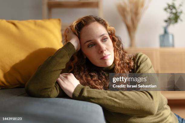 portrait of a young woman with long curly red hair sitting on the floor and leaning on a sofa, looking away with sad face. moment of sadness and worry in the living room of her house. - concerned woman stock-fotos und bilder