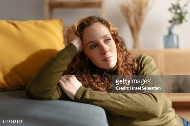portrait of a young woman with long curly red hair sitting on the floor and leaning on a sofa, looking away with sad face. moment of sadness and worry in the living room of her house. - donna triste foto e immagini stock