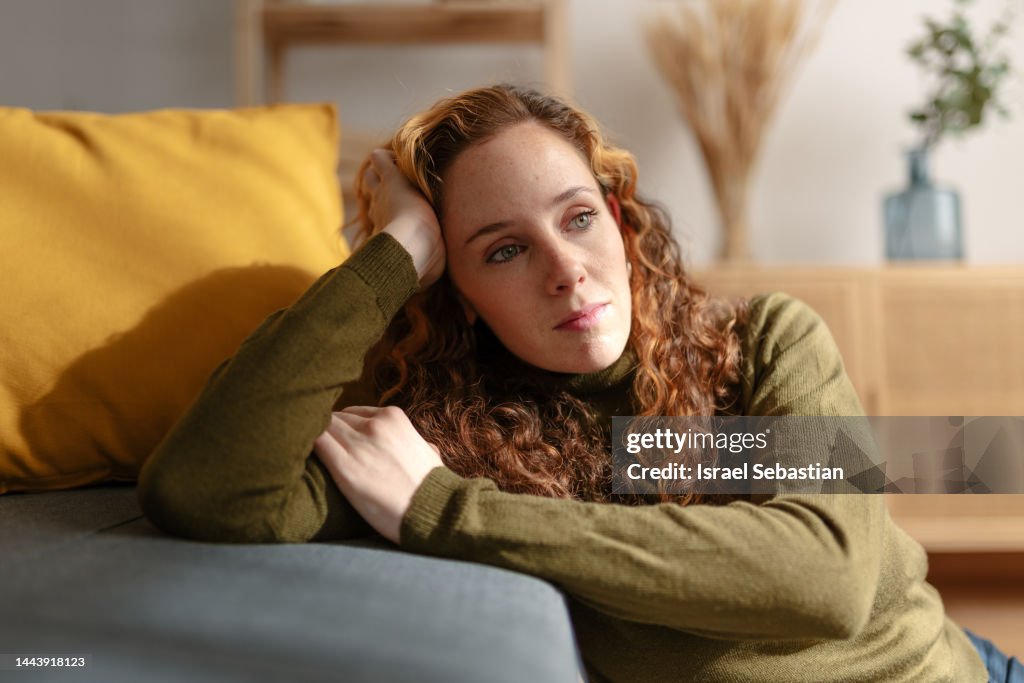 Portrait of a young woman with long curly red hair sitting on the floor and leaning on a sofa, looking away with sad face. Moment of sadness and worry in the living room of her house.