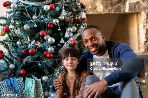 adopted latin american little girl and black daddy sitting in front of the christmas tree facing camera smiling - latin beauty stockfoto's en -beelden