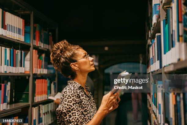 female university student choosing book from shelf in library - bibliotheek stockfoto's en -beelden