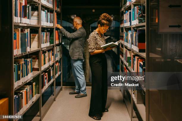 female student reading book by man in library at college - bookshelf photos et images de collection