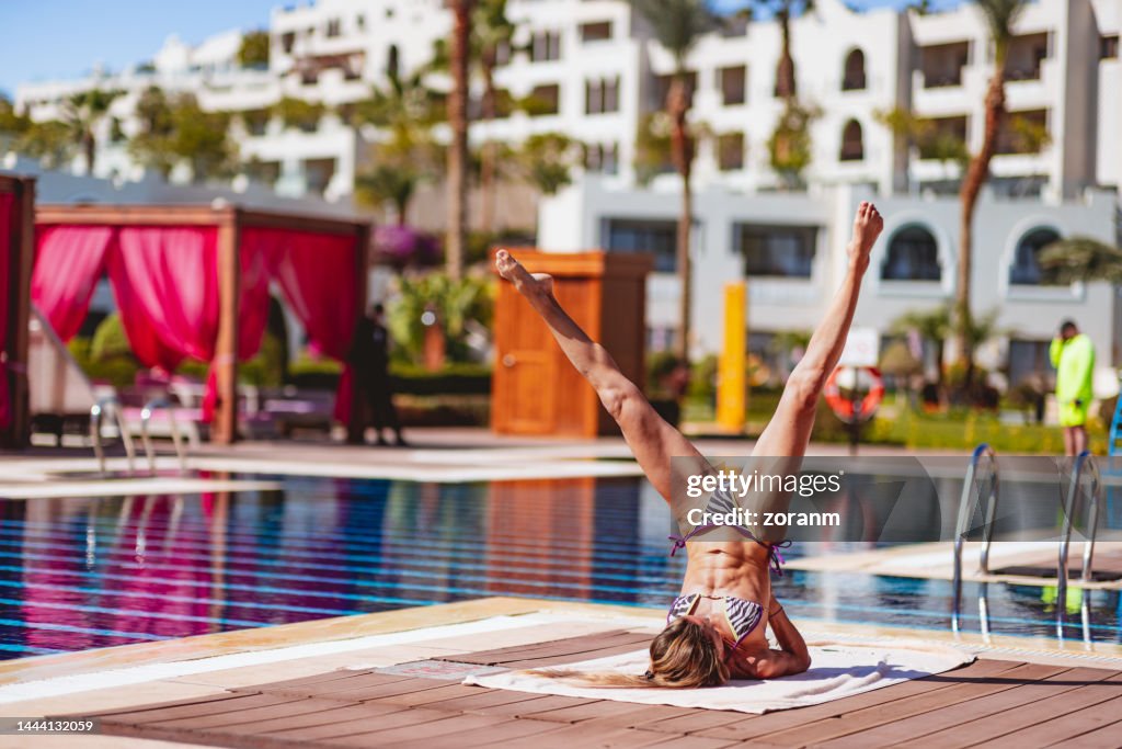 Mid adult woman exercising at poolside on mat, assuming pose for balance in shoulder stand