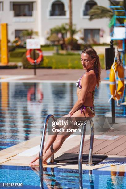 beautiful mid adult woman in bikini leaning on rails by the pool and sunbathing - ladder leaning stock pictures, royalty-free photos & images