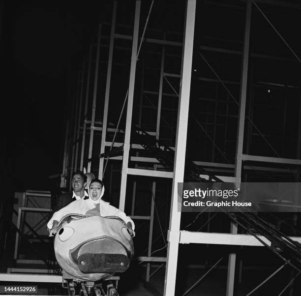 Northern Irish actor Maxwell Reed and his wife, British actress Joan Collins wearing a white headscarf, both sitting in the car of an roller coaster,...
