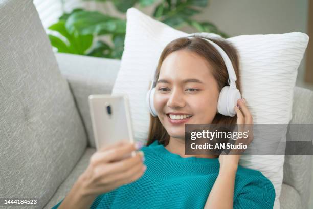 beautiful young woman with headphones relaxing on sofa. female listening to music using smart phone. chill out and leisure concept - smartphone video stock pictures, royalty-free photos & images