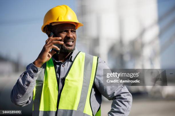 worried male engineer with hardhat talking on mobile phone at industrial facility - frustrated workman stock pictures, royalty-free photos & images