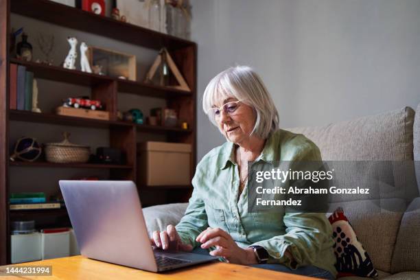 senior woman using a laptop sitting on the sofa in a house - retirement fotografías e imágenes de stock