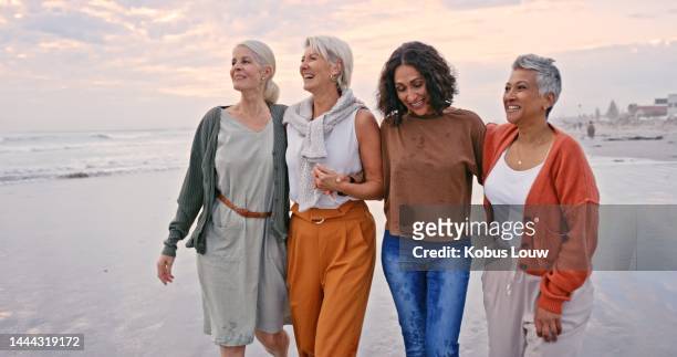 spiaggia, amici e vacanze con un gruppo di donne senior che camminano sulla sabbia in riva al mare o all'oceano. natura, acqua e amicizia con la diversità e femmine mature che ridono al tramonto nel suo pensionamento - donne mature foto e immagini stock