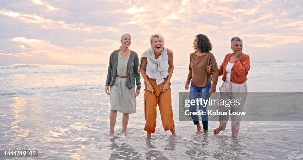 happy mature women in group at beach in sunset, laughing at joke and and feet in water together. happiness, smile and friendship in retirement, girl friends having fun on ocean holiday spending time. - mature woman in water stock pictures, royalty-free photos & images